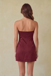 A woman viewed from the back wearing a strapless mini dress in a deep burgundy color, set against a textured beige background.