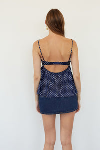 A woman viewed from the back wearing a navy blue spaghetti-strap top with white polka dots and a denim mini skirt against a plain white background.
