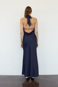A woman viewed from the back wearing a navy blue satin halter maxi dress with a cowl neck and open back, standing against a plain white wall on a dark wood floor.