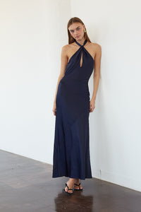 A woman modeling a navy blue halter-neck maxi dress with a keyhole cutout at the chest, standing against a white wall on a dark wood floor.