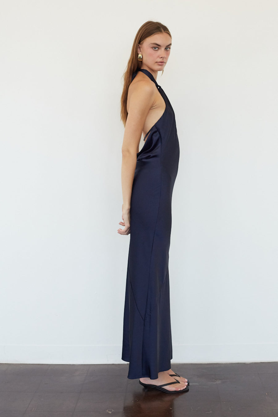 A woman modeling a sleeveless, halter-neck maxi dress in dark navy blue, standing in profile against a plain white wall.
