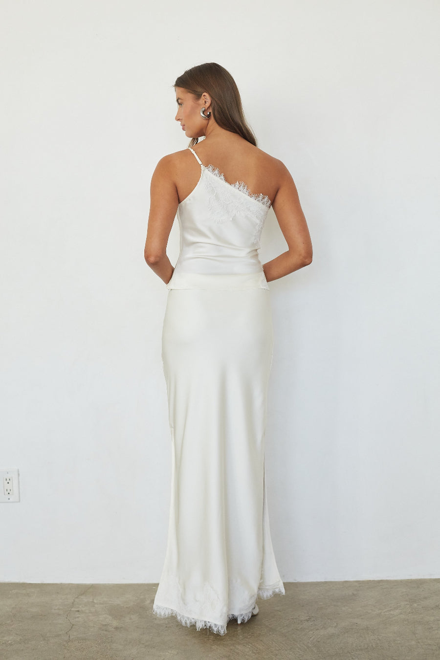 brunette model wearing a white lace trim asymmetrical top and satin skirt back image