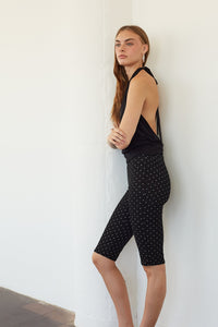 A woman wearing a black halter top and black biker shorts with small white polka dots, leaning against a white wall in a minimalist indoor setting.
