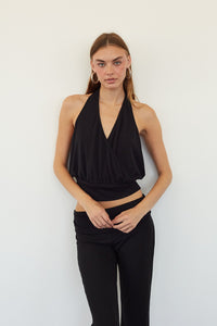 A woman modeling a black halter wrap-style crop top and black pants against a plain white wall.
