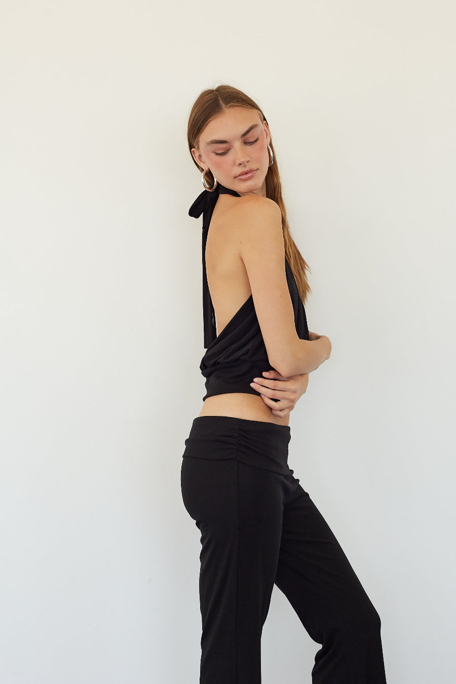 A woman modeling a black halter crop top with a draped back and black pants, standing against a plain white wall.