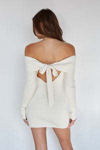 Woman wearing a white off-shoulder sweater with a bow detail on a plain background