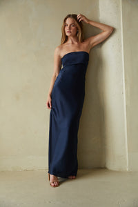 A woman wearing a navy blue pleated satin maxi dress with a strapless neckline, standing in a room with textured walls.