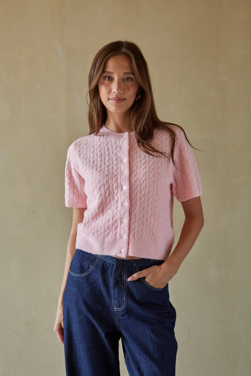 Woman wearing a pink knitted cardigan and blue pants against a beige background