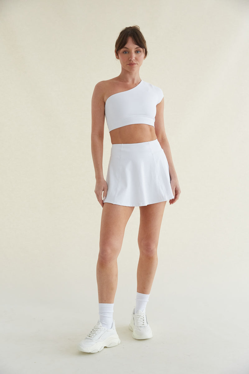 front view cream seamless background with a model wearing a one shoulder cropped athleisure top and a matching flared mini skort with pockets - crew socks and chunky dad sneakers - women's athleisure 